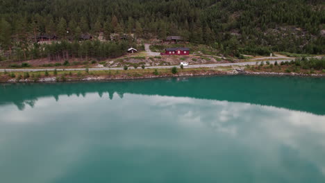 Stunning-Aerial-View-of-a-Country-Road-Lining-the-Coast-of-a-Teal,-Green,-Turquoise-Lake,-The-Drone-is-Following-some-Cars,-Norway,-Scandinavia