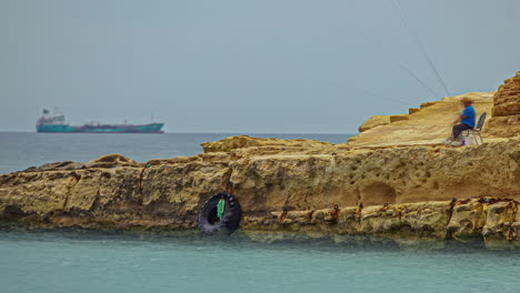 fisherman sitting on a chair on rock formation, catching fish with fishing rod in daytime