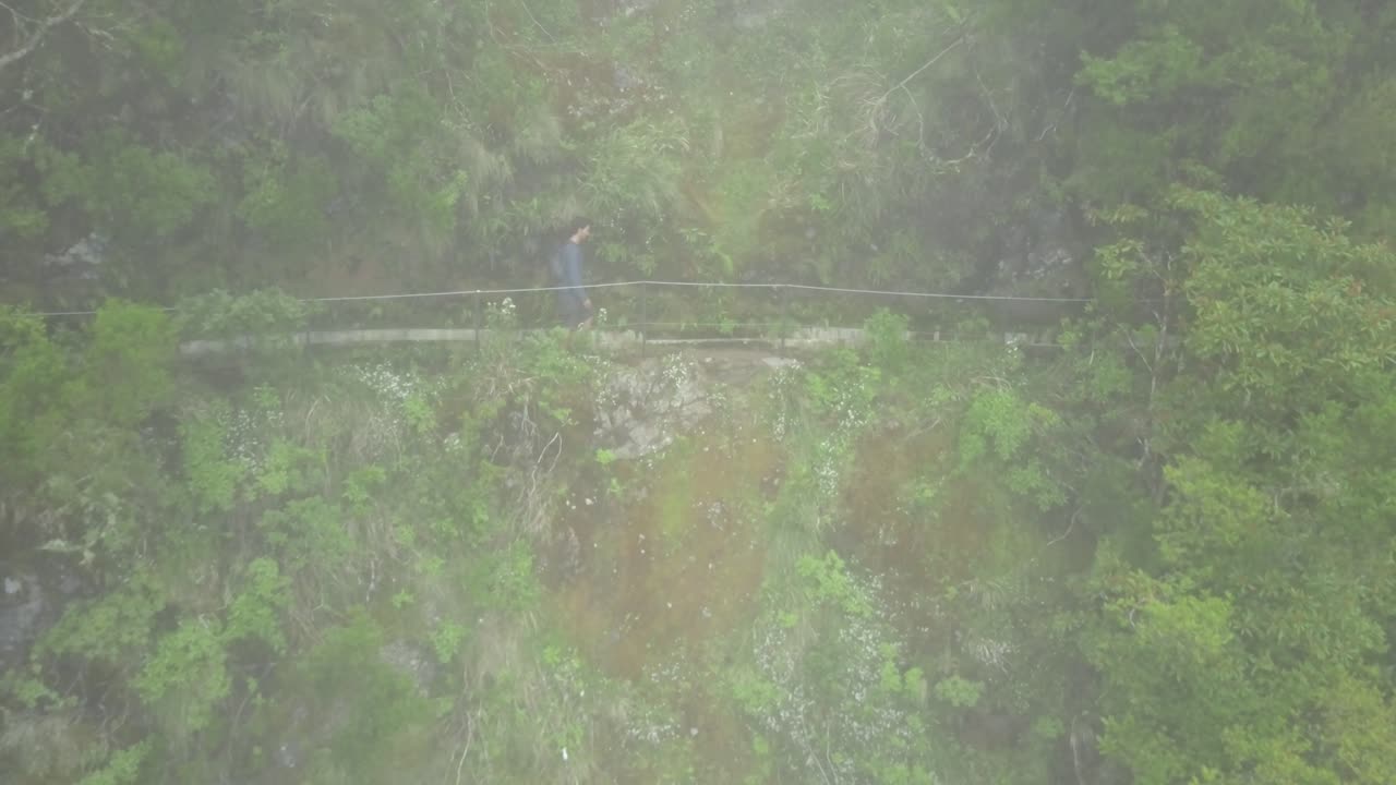 Madeira, Portugal - Male Backpacker Walking At The Trail On The Side Of The Lush Mountain On A Foggy Day - Aerial Drone Shot