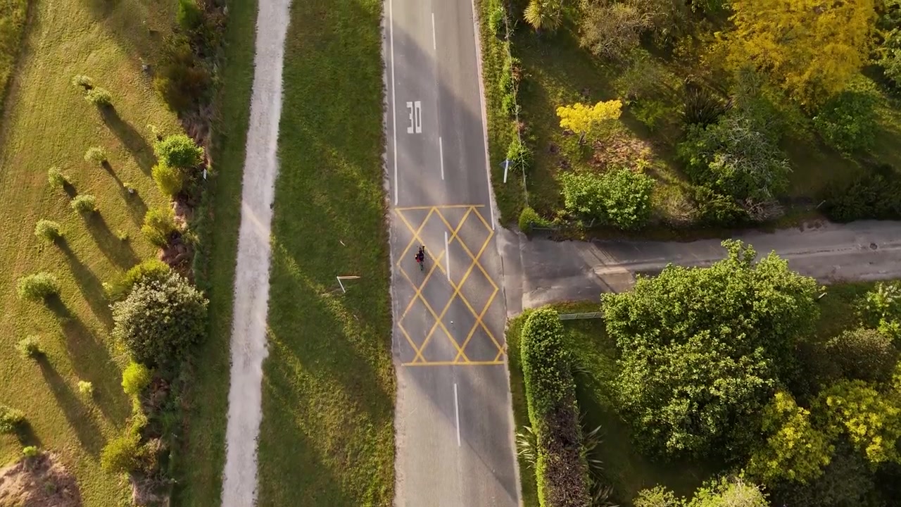 Ciclista che pedala lungo una strada tranquilla nella regione di Marahau Abel Tasman in Nuova Zelanda, circondato da un paesaggio verde e lunghe ombre del tramonto. Momento di viaggio panoramico e pacifico. Ripresa aerea con drone dall'alto che segue.