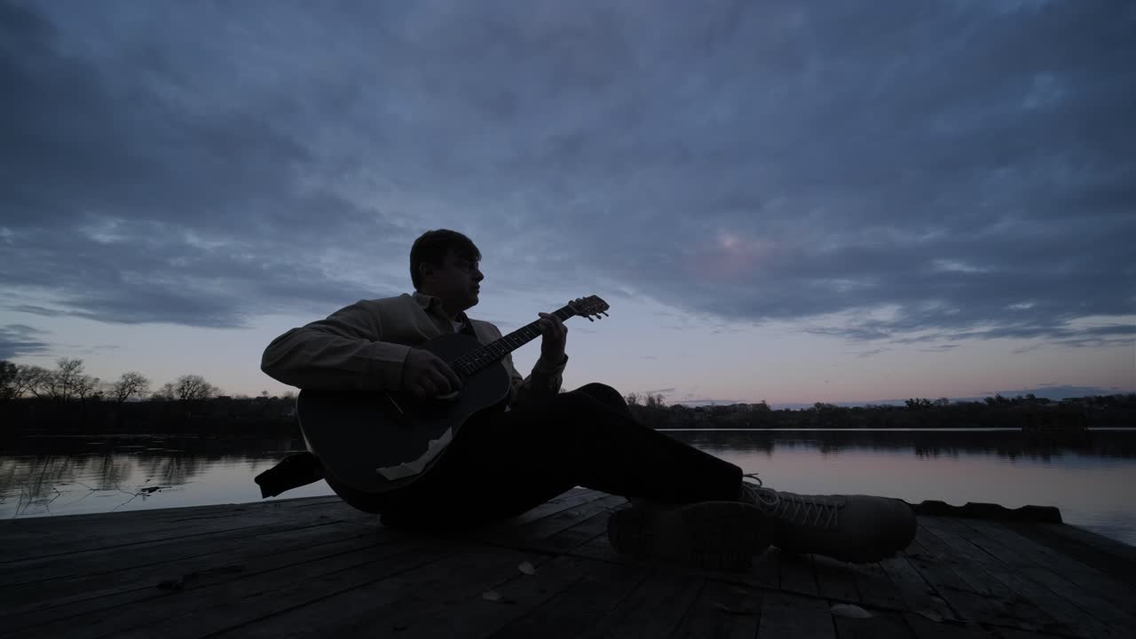 silueta de músico en negro tocando la guitarra sentado en el terraplén del muelle al atardecer