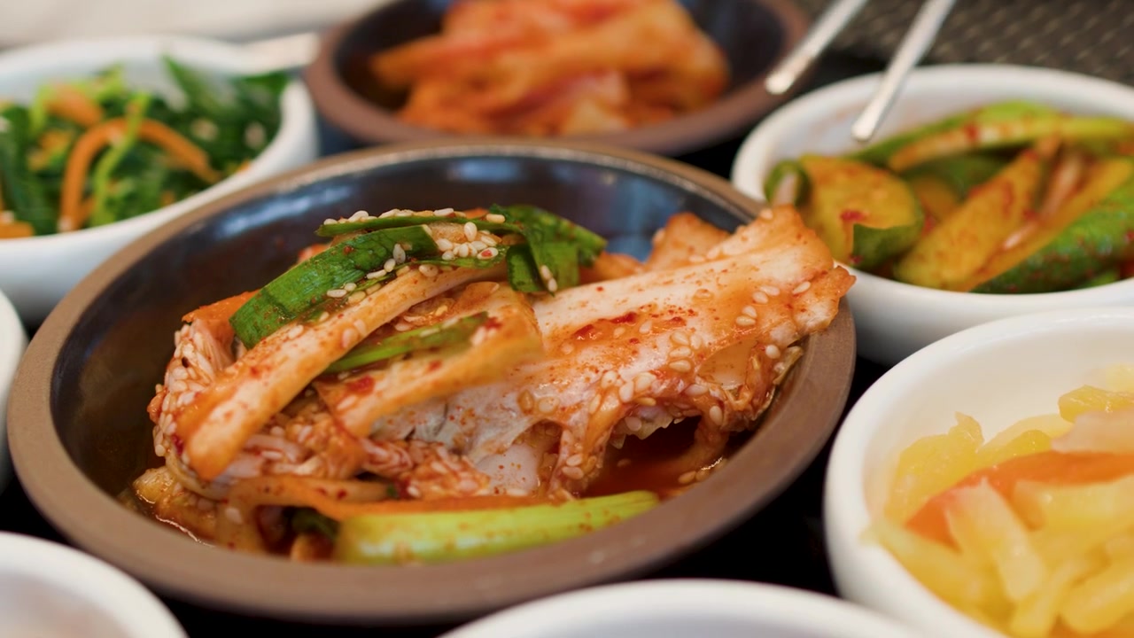 Bidikan close-up kimchi sawi pedas dan aneka banchan dengan sumpit logam yang sedang digunakan