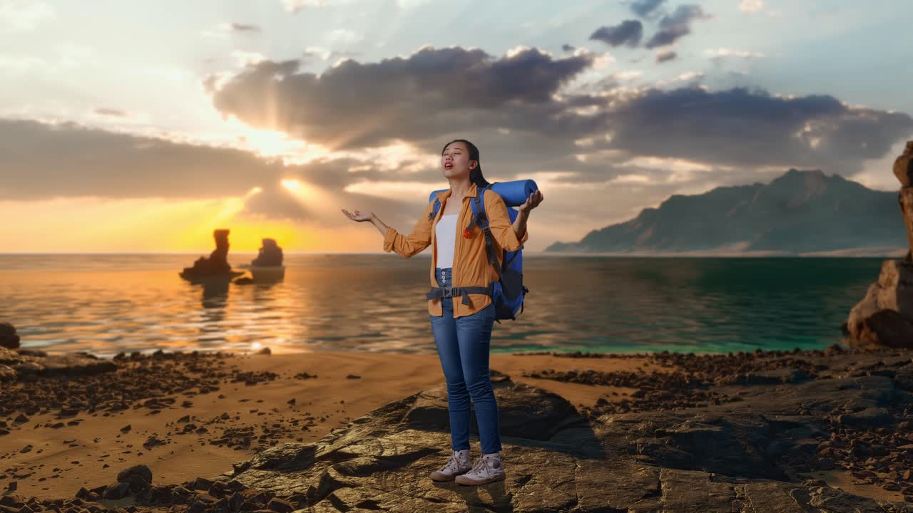 Inquadratura laterale a figura intera di un'escursionista asiatica con zaino da alpinismo che sorride e allarga le braccia, godendosi la vista mentre si trova in spiaggia al tramonto