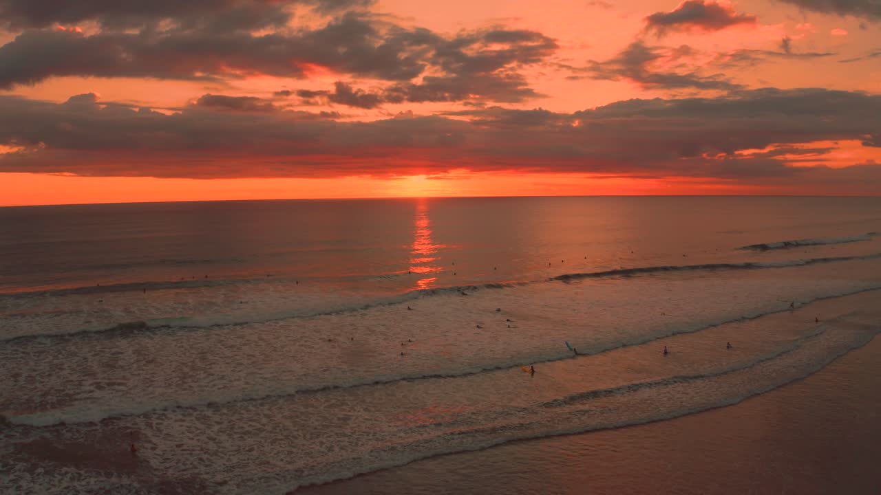 Epic aerial shot of a wide sandy beach at the Pacific Coast in Costa Rica at sunset with a beautiful colored sky and clouds above the ocean. Surfers are waiting for the perfect wave. Shot in 4K.
