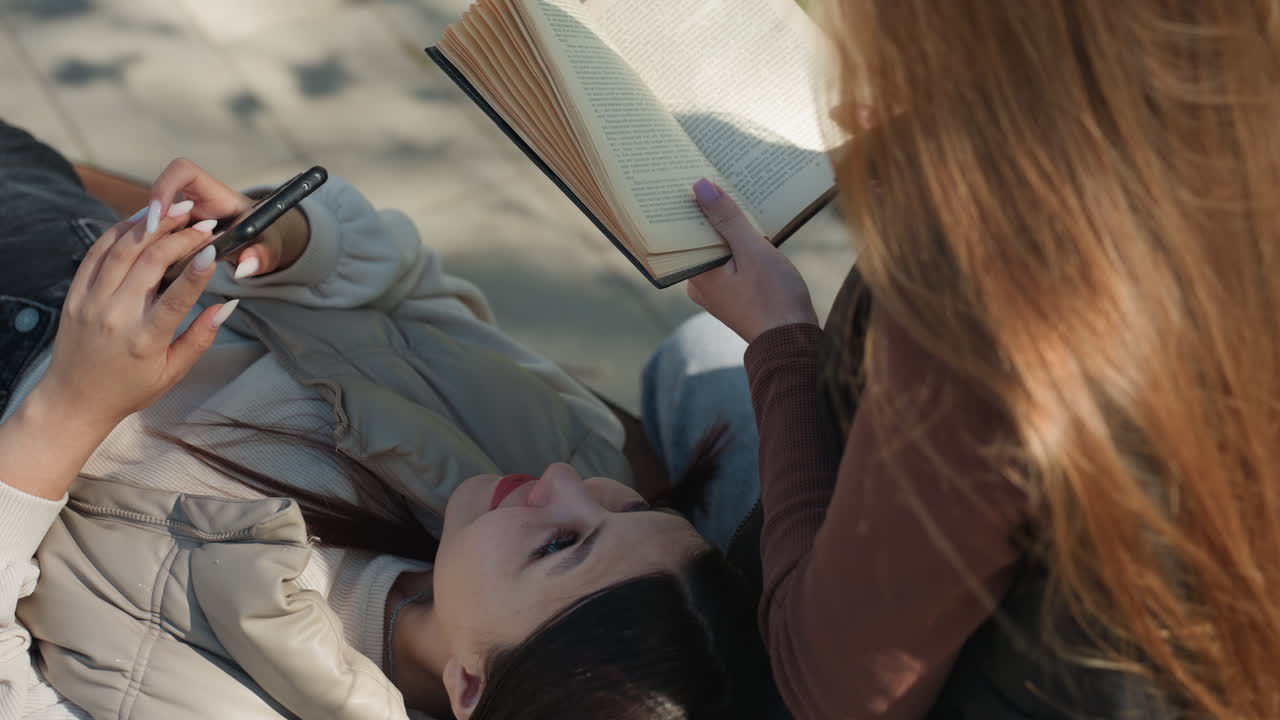 Белая девушка с длинными волосами сидит и перелистывает страницы книги, а ее подруга азиатской внешности проверяет телефон. Снято сверху, уютный перерыв на учебу с очками и общими медиа; общая атмосфера кампуса и игривый обмен