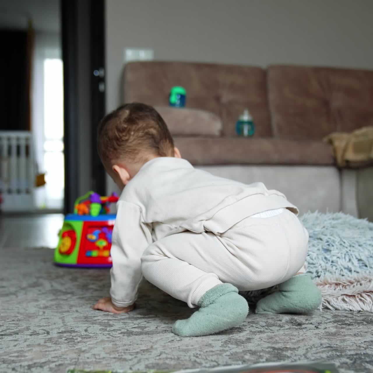 Rear view of a baby in white suit sitting on the floor and eating a cookie. Kid drops the food on the floor, stands up to get it back and eat