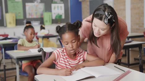 happy diverse female teacher helping schoolgirl at desk in elementary school class, slow motion