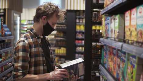 male customer in the supermarket during pandemic time. man reading the label on the pack of pasta. shelves and department row on the background. wearing black protective mask and gloves