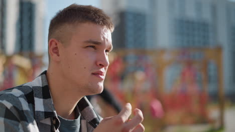 close up of man in checkered shirt takes red medicine outdoors with focused expression while kids ride scooter in colorful blurred background featuring urban buildings and wooden pergola structure
