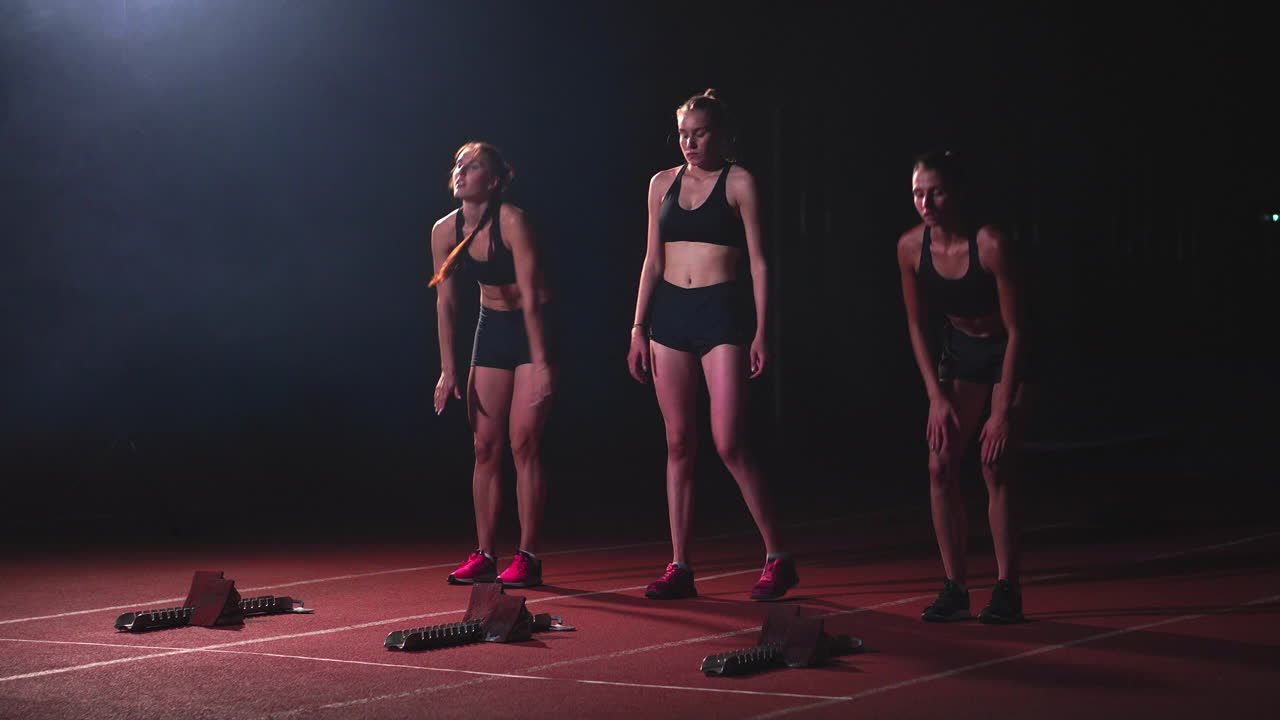 트레드밀에 밤에 운동 선수의 검은 옷을 입은 세 명의 스포츠 소녀는 앉은 위치에서 스프린트 거리에서 경주를 시작할 것입니다.