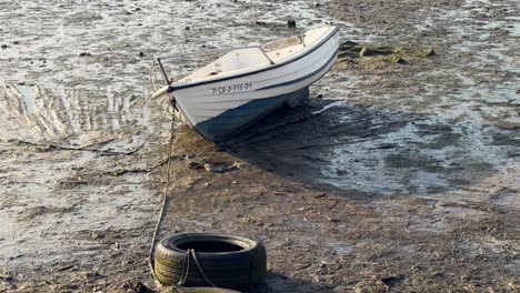 Walking-near-traditional-fishing-boat-on-the-sand,-dried-up-and-muddied-ground-surface,-old-fishing-boat-and-tires-at-river-empty-bottom