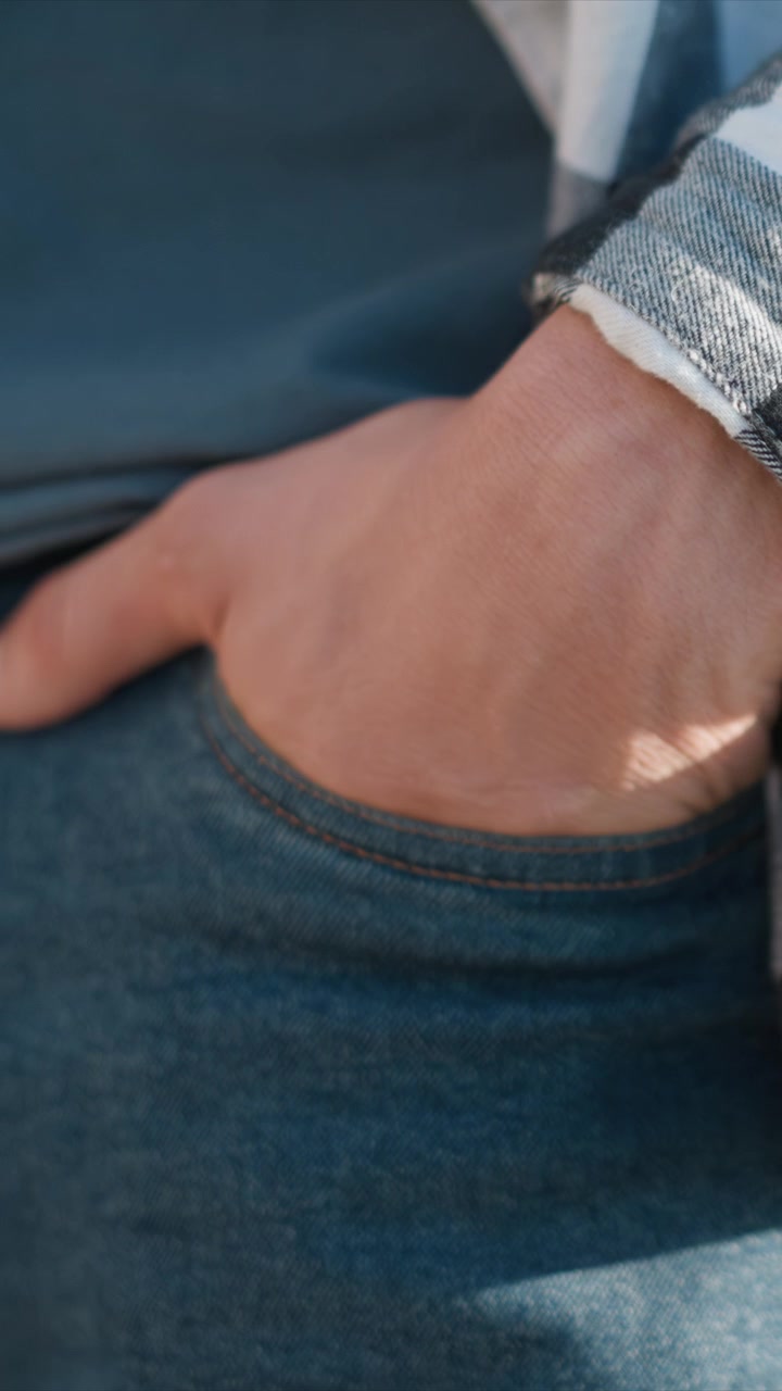 Primo piano della mano di un uomo che riposa nella tasca di jeans blu con cuciture visibili e texture del tessuto. La scena mantiene un'illuminazione soffusa e costante, concentrandosi sul materiale denim dettagliato.