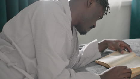 man in white robe reading large book while lying on bed, deeply focused on pages, soft light streaming from window creating peaceful, serene atmosphere, deep in thought and contemplation