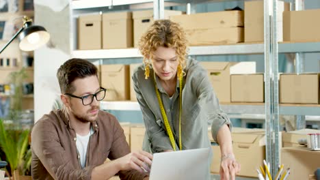 caucasian young professional designers working and speaking while browsing on laptop computer in clothing shop warehouse