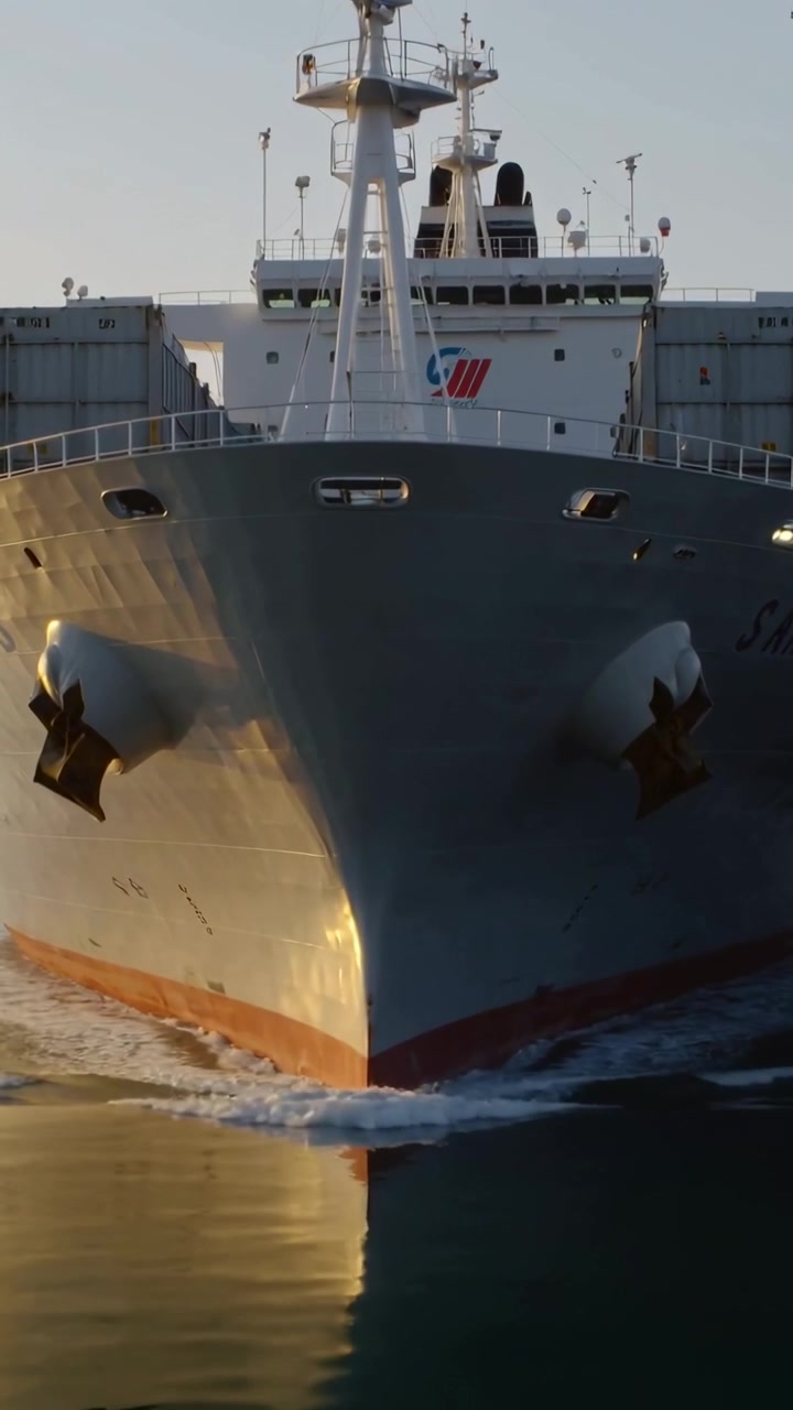 Vertical video: Engine powering cargo ship passing camera through calm port channel with containers
