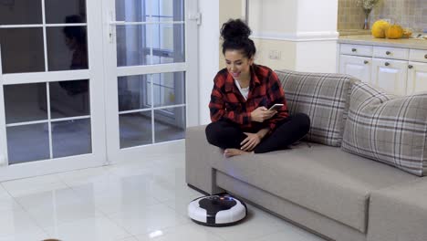 young woman relaxes with phone while the vacuum-cleaning robot clean the floor