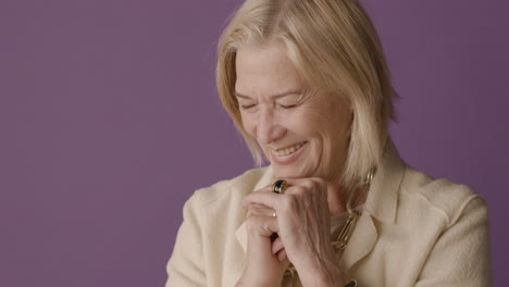 Blonde-mature-woman-with-blue-eyes-dressed-in-jacket-and-accessories-posing-with-fists-on-chin-and-smiling-with-eyes-closed-on-purple-background