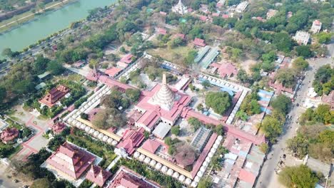aerial perspective of mandalay, myanmar, highlighting a sprawling pagoda complex with intricate architecture, lush greenery, and surrounding urban streets near a serene riverbank