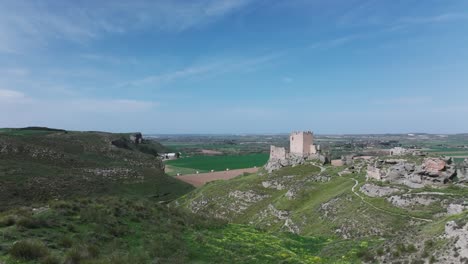 drone flight taking off from a platform viewing the 9th century oreja castle and the remains of its walls behind in an intense green there are crop fields with a blue sky in ontigola toledo spain