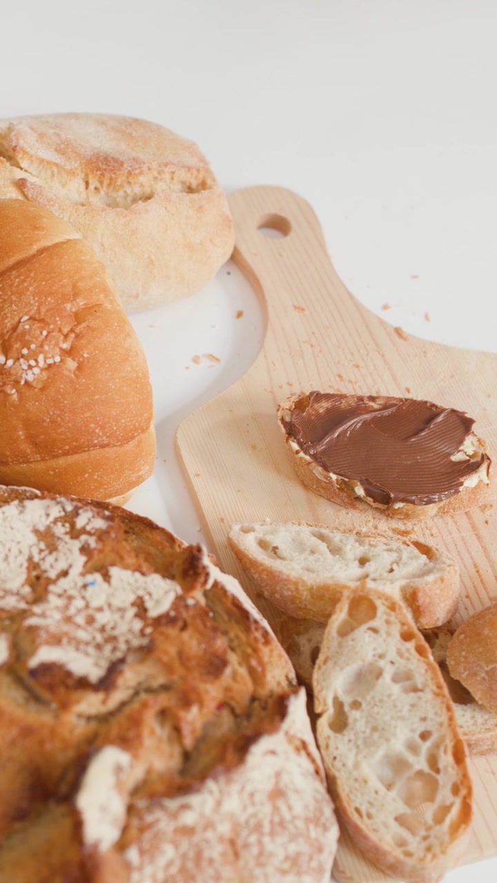 Close-up shot of hands spreading chocolate cream on bread amidst various bakery pastries.