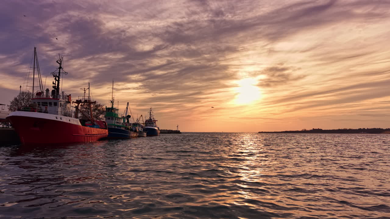 Un vivace tramonto si affaccia sulle barche da pesca allineate lungo le acque serene del porto, proiettando riflessi sulle onde leggere durante le ore del crepuscolo.
