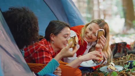 group of female friends on camping holiday in forest lying in tent eating s'mores