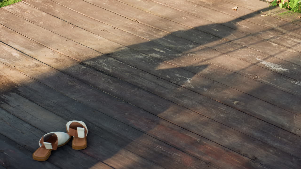 Sunlit Wooden Deck With Abandoned Sandals, Looming Human Silhouette Crosses Floorboards, Long Late Afternoon Shadow, Warm Tones, Weathered Planks, Quiet Solitude, Cinematic Suspense