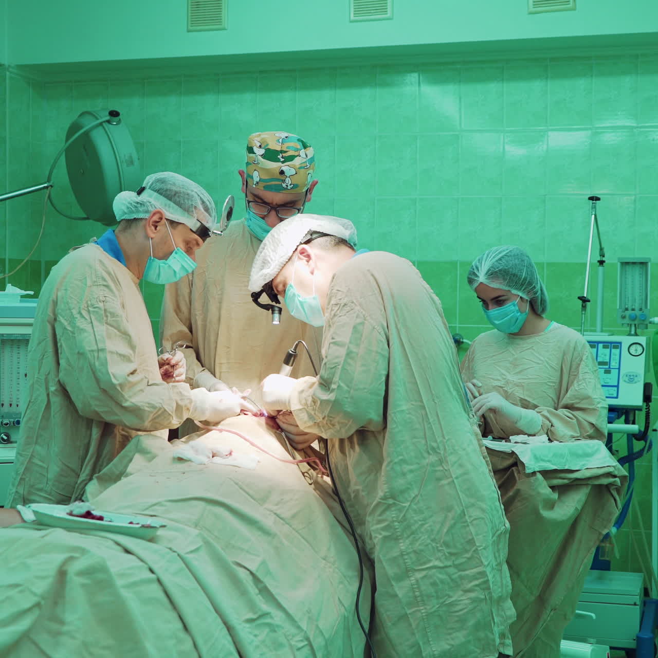 Teamwork of professional surgeons in the operating room. Doctors in special uniform work with medical equipment during the operation on a patient on the green background in clinic.