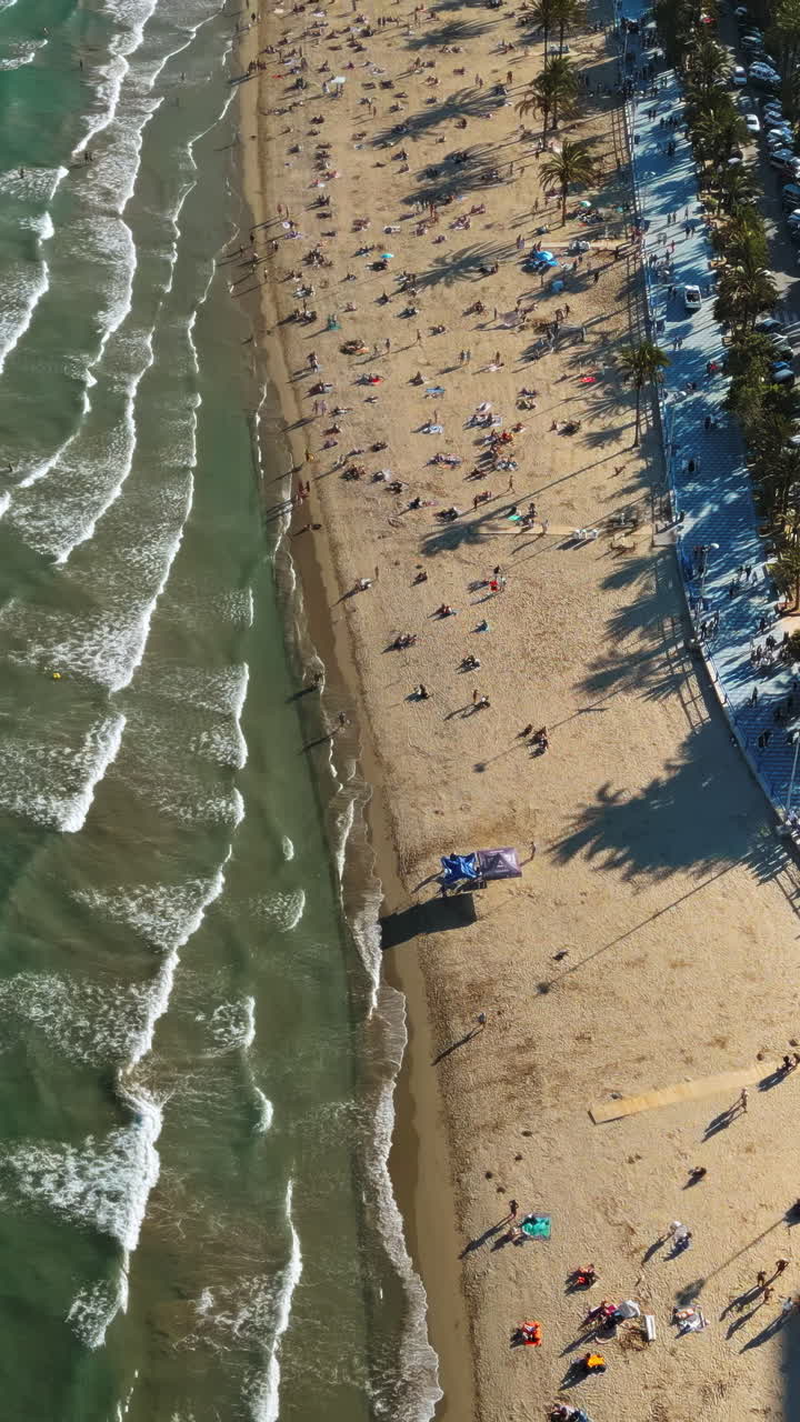 Aerial drone view of cars moving along the coastline with people relaxing on the beach in Alicante, Spain in daylight. Vertical