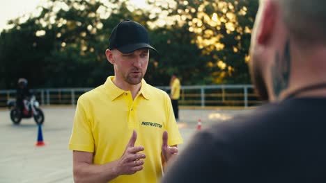 a man instructor in a yellow t-shirt and cap communicates with a motorcycle driving student at the training ground of a motorcycle school