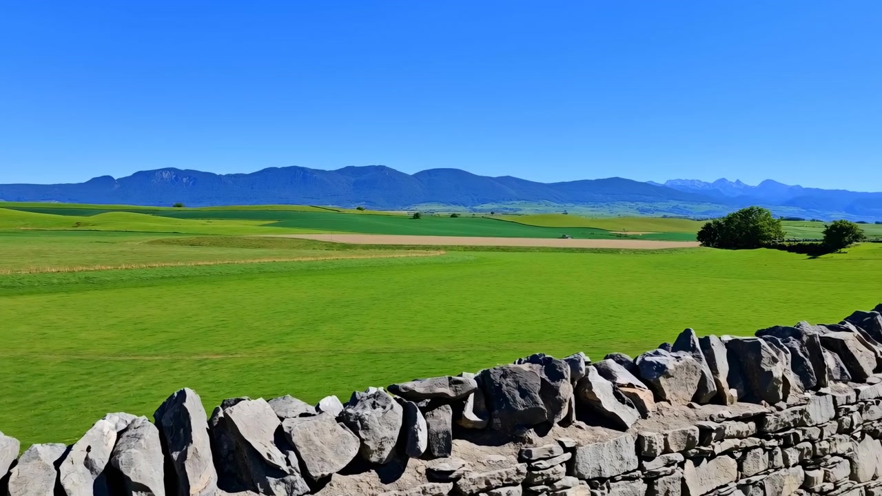 Campi verdi soleggiati oltre un muro di pietra, dolci colline e lontane montagne blu sotto un cielo sereno, vibrante.