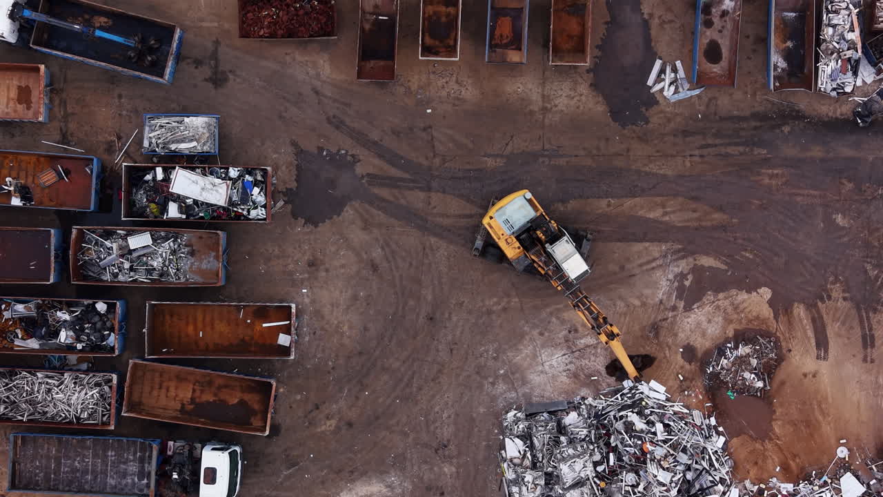 Vista dall'alto di un deposito di rottami metallici con un escavatore giallo che smista i rottami tra file di container scarrabili arrugginiti pieni di rifiuti metallici misti.