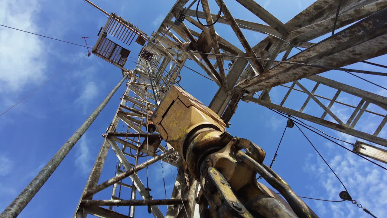 Borer vibrates as it drills ground. Footage at the base of a high derrick at the site for production oil. Low angle view.