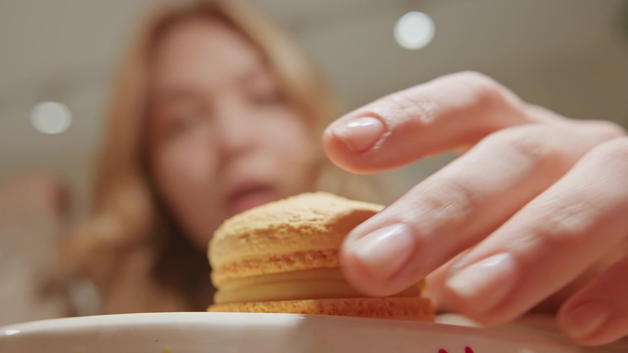 Close up of woman reaching for and picking up orange macaron from white plate. subject hand with neatly manicured nails interacts directly with dessert emphasizing texture detail. Soft