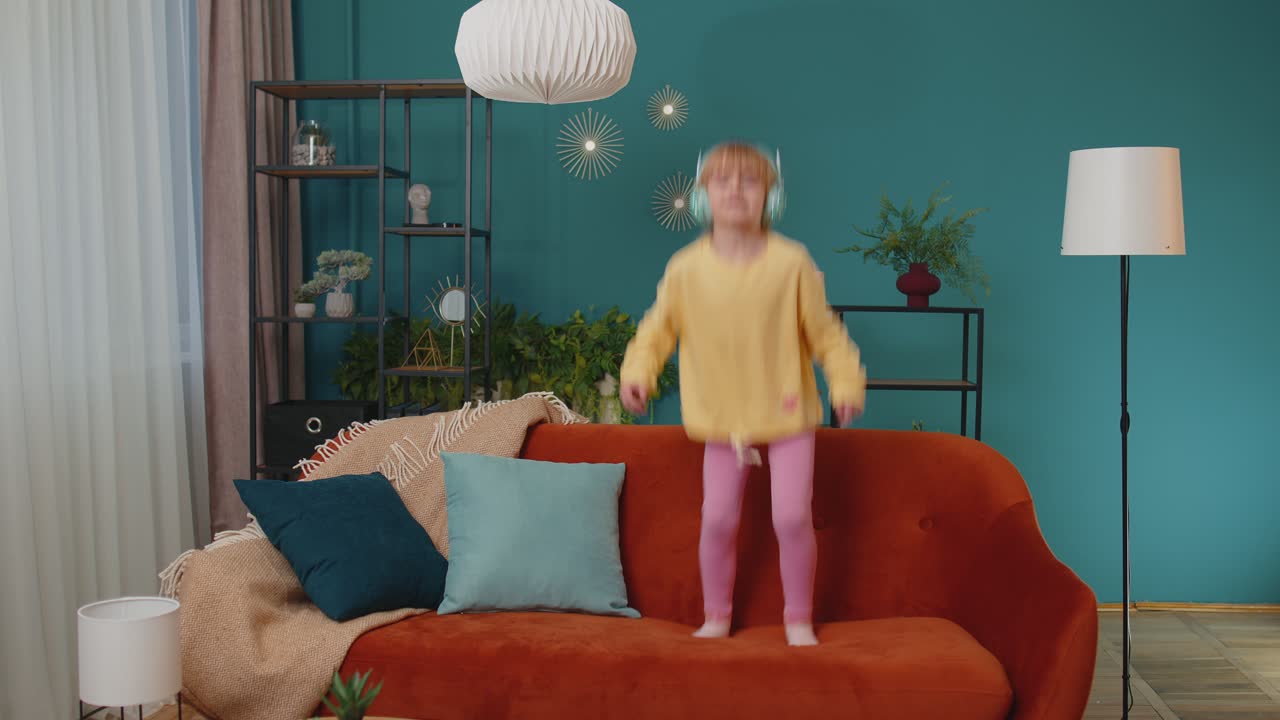 Happy child girl kid in headphones dancing and jumping while listening to the music at home alone
