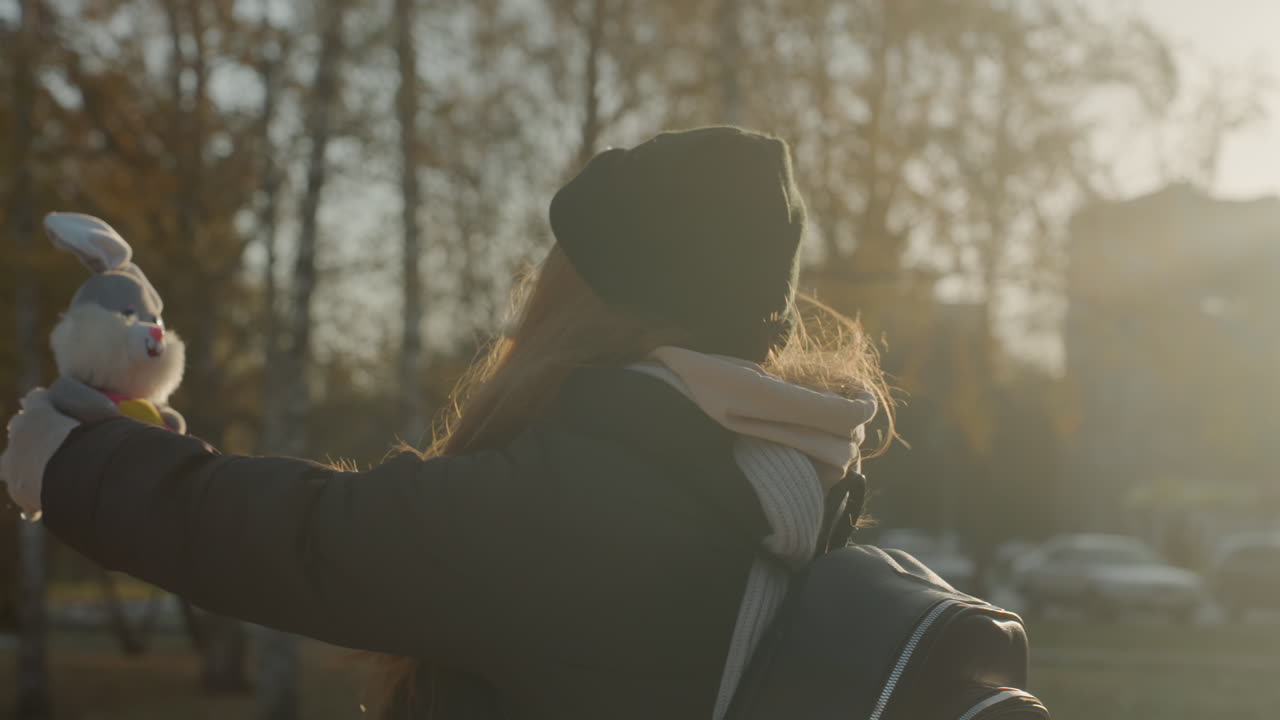 Young woman offers stuffed toy in autumn park at golden hour, backlit profile wearing beanie, scarf and backpack, smiling while extending arms, soft sunlight creating lens flare, candid warm moment