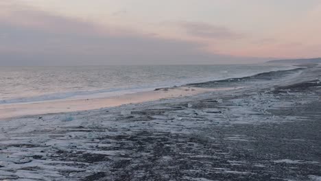 aerial landscape view over ocean waves crashing on to diamond beach, covered in snow, at sunset