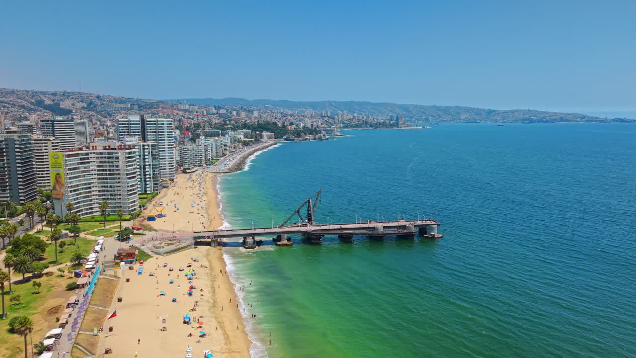 Ripresa aerea con carrello (dolly in) che mostra il Molo di Vergara con la Spiaggia El Sol a Viña del Mar in una giornata di sole con traffico intenso e una strada costiera con molta vegetazione, Cile