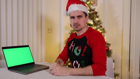 hombre con gorro de papá noel y suéter mirando la cámara apuntando a la pantalla verde de la tecla croma portátil con expresiones de juicio infelices