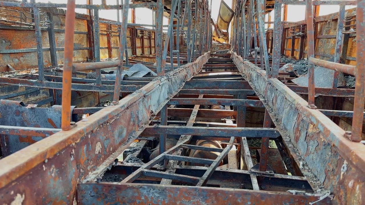 Tracking view through charred sleeper bus floor structure with exposed metal framework, rusted surfaces and severe fire destruction inside damaged vehicle!