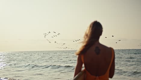 Rear-view-of-a-blonde-girl-in-an-orange-swimsuit-standing-with-a-surfboard-on-the-seashore-and-looking-at-the-sea-and-the-sky-where-a-large-flock-of-birds-are-flying-at-Sunrise-in-the-morning