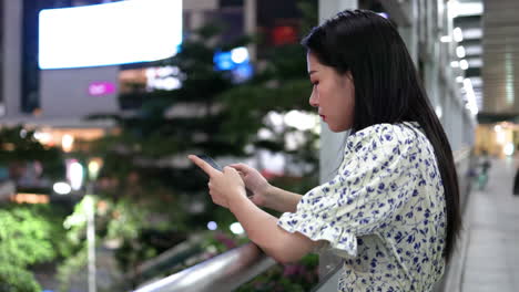 portrait of lonely girl leaning on the railing of overpass playing with her mobile phone at night, cute sad woman being depressed