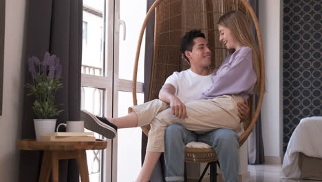 portrait of young couple looking at each other while sitting in a wicker chair together at home