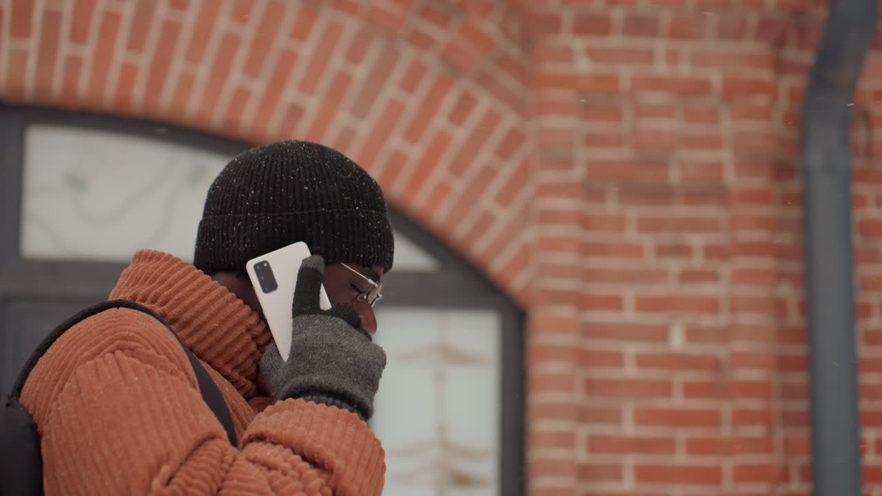 Homem negro ao telefone na neve, close-up perto de um edifício de tijolos mostrando um passageiro focado fazendo uma chamada urgente, primeiro ângulo retrata o passageiro verificando os horários de trânsito, segundo ângulo mostra o arquiteto negociando alvenaria