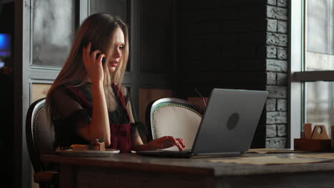 mujer joven sentada en una cafetería en una mesa de madera, bebiendo café y usando un teléfono inteligente. en la mesa hay una computadora portátil. chica navegando por internet, chateando, blogueando. mujer sosteniendo el teléfono y mirando su pantalla.