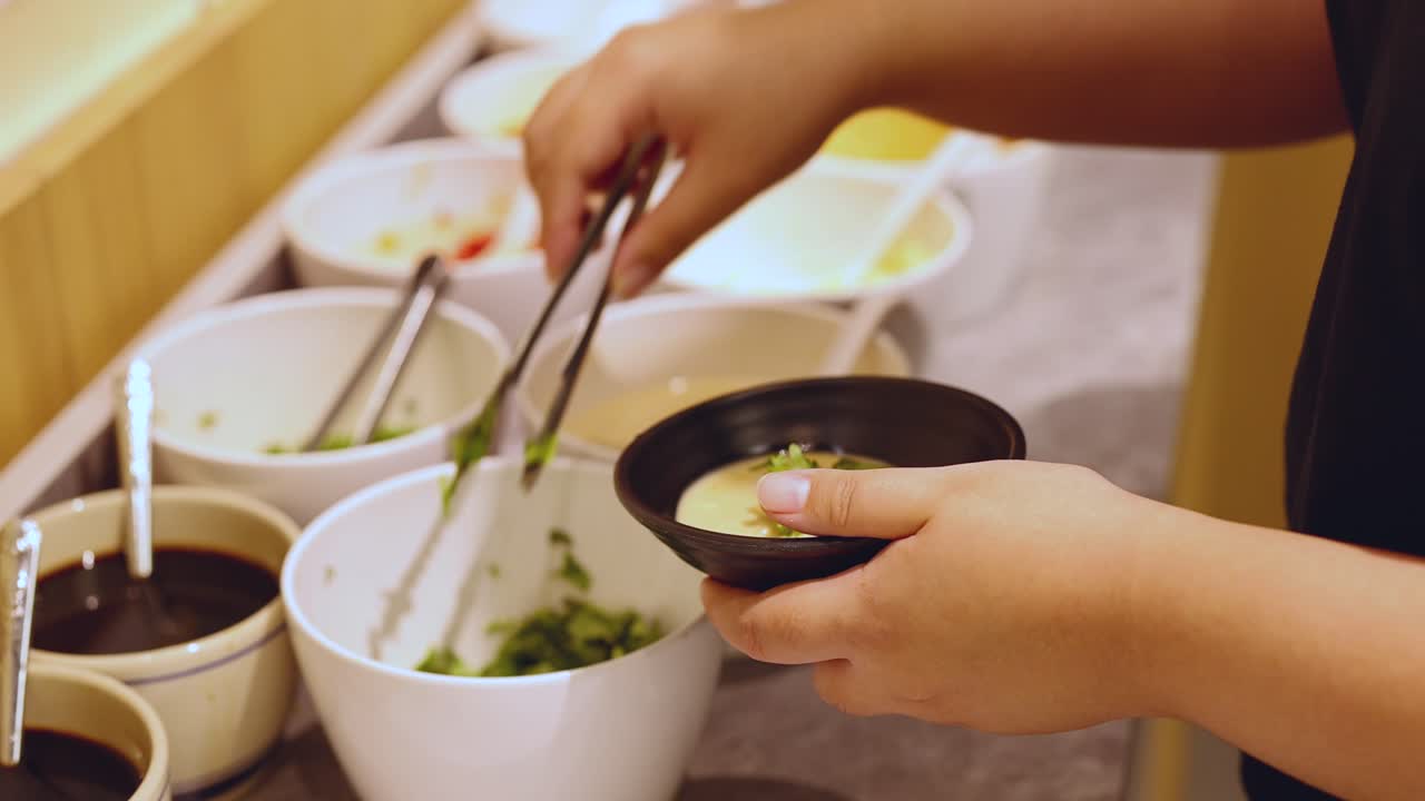 Person uses tongs to add condiments to bowl at self-service hot pot sauce station