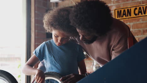 Close-up-of-pre-teen-mixed-race-boy-using-a-spanner-to-attach-the-wheel-of-his-racing-kart-at-a-workbench-in-the-garage-with-his-father-standing-behind-him-helping,-selective-focus