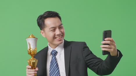 close up of asian business man in a suit and tie with a gold medal holding a gold trophy and taking photo on smartphone as the first winner on green screen background in the studio