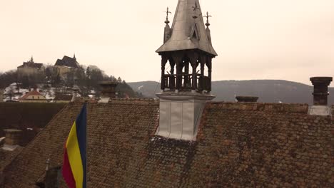 a rotating drone shot, capturing the rooftop of a vintage architechture in the city of sighisoara on an afternoon with a community flag hoisted beside and a cityscape in the background