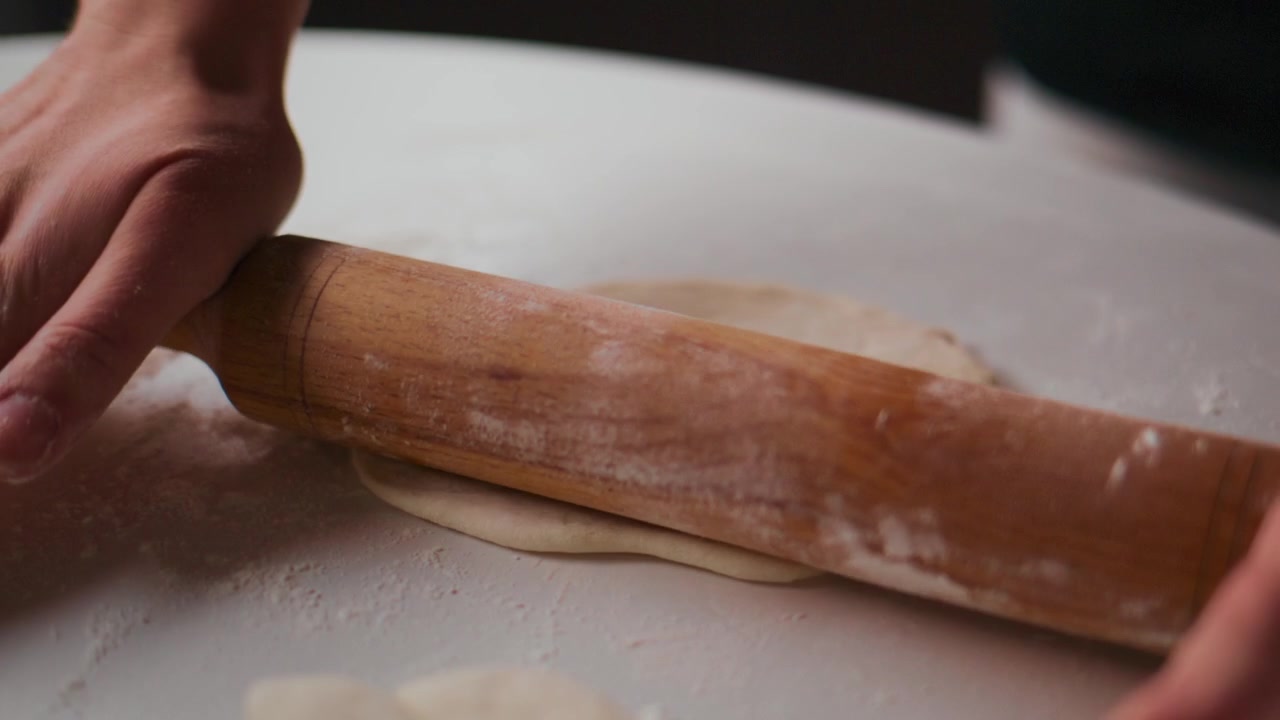 Primo piano di mani che stendono l'impasto con un mattarello di legno su una superficie infarinata. Persona concentrata, movimento attento e preciso. Ideale per progetti di cucina, pasticceria o preparazione alimentare. Adatto per ricette, tutorial.